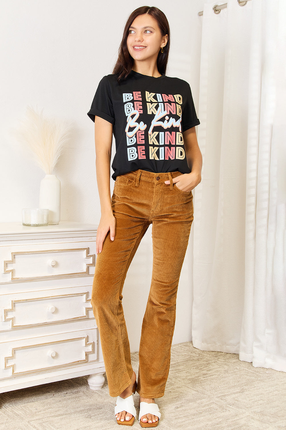 Woman wearing Simply Love BE KIND graphic round neck T-shirt with brown pants, showcasing casual, stylish outfit.