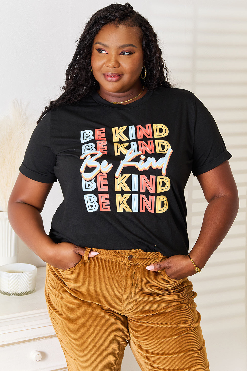 Woman wearing a black "BE KIND" graphic round neck t-shirt with colorful print, paired with mustard pants, posing indoors.