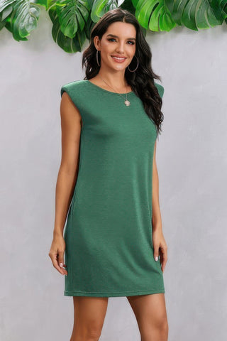 Woman in emerald green sleeveless dress with hoop earrings and necklace, standing against white wall with tropical plants in background, smiling...