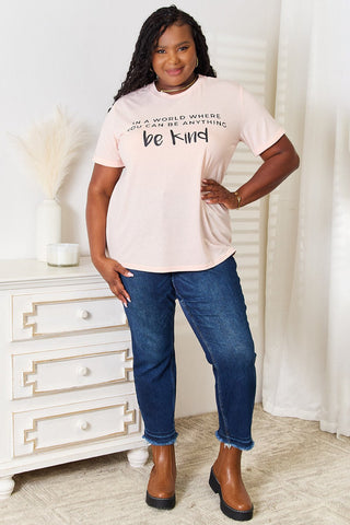 Woman wearing slogan graphic cuffed t-shirt with "be kind" message, paired with blue jeans and brown shoes in a stylish indoor setting.