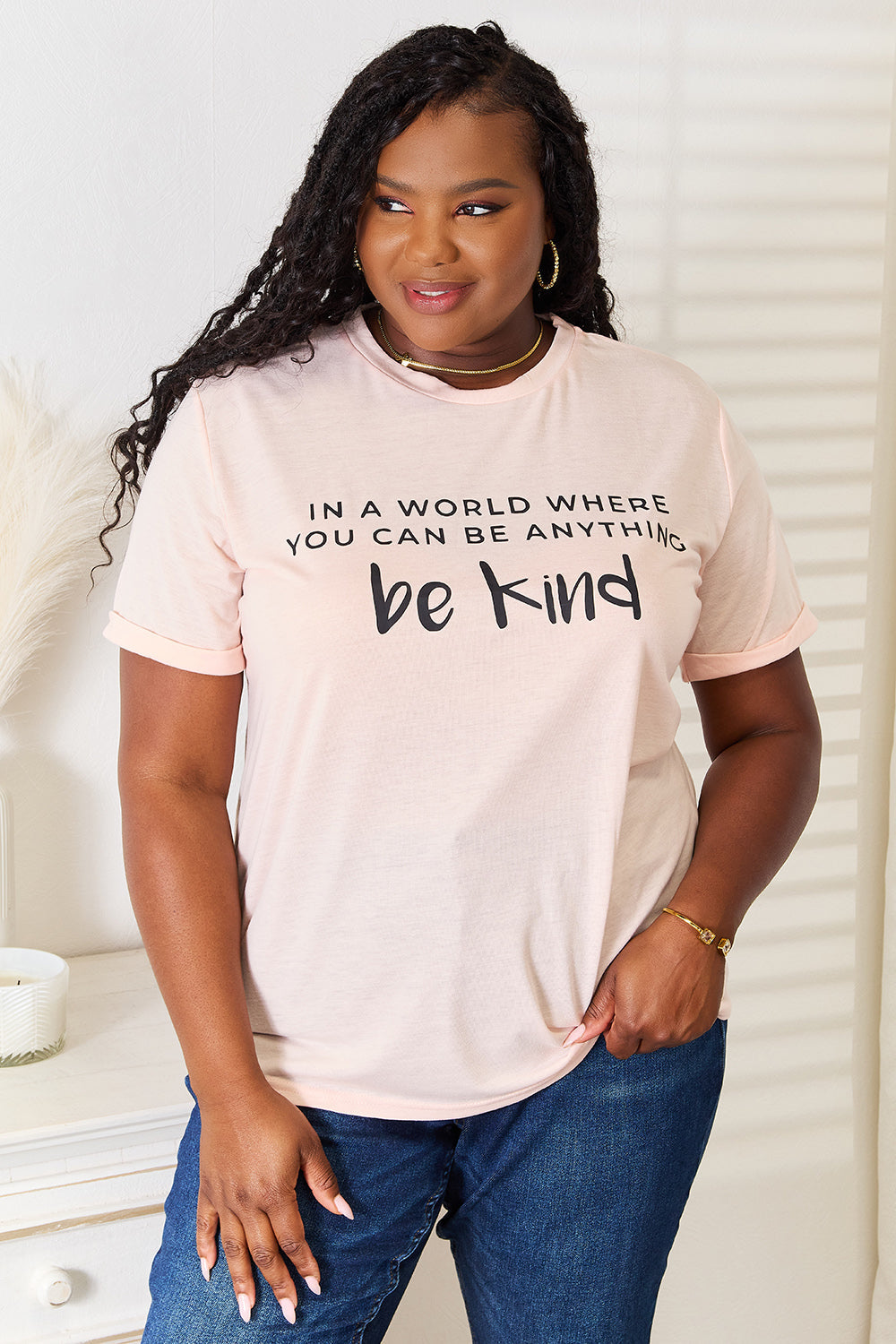 Woman wearing a pink slogan graphic cuffed T-shirt with "Be Kind" text, paired with blue jeans.