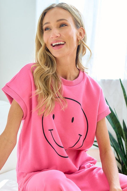 Woman wearing pink smiley face t-shirt with drop shoulder, sitting and smiling in a bright room.