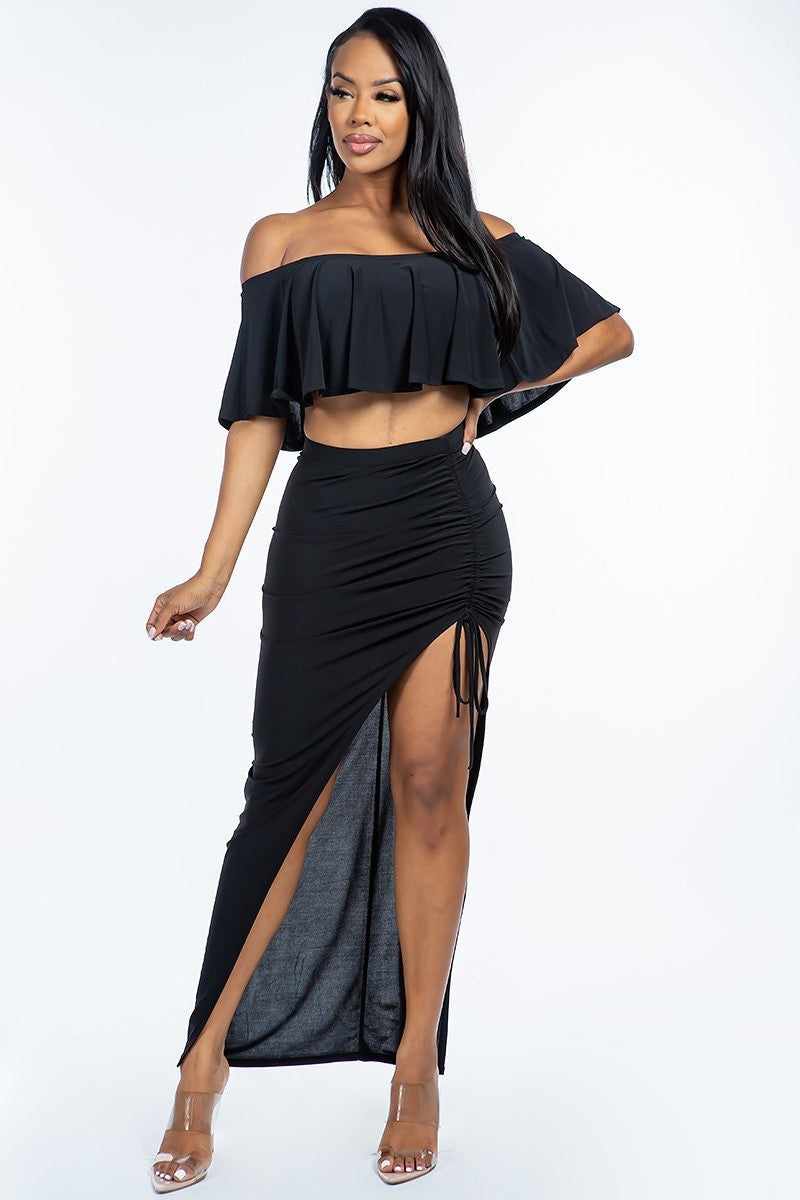 Woman wearing black off-the-shoulder cropped top and ruched maxi skirt set with slit, made from polyester and spandex.