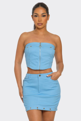 Woman in light blue two-piece outfit consisting of strapless crop top with front zipper and matching mini skirt with metal studs, standing against ...