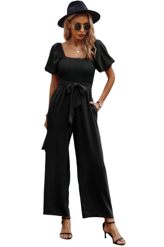 Woman in black wide-leg jumpsuit with puffy sleeves and waist tie, wearing black fedora hat and sunglasses, posing against white background with ha...