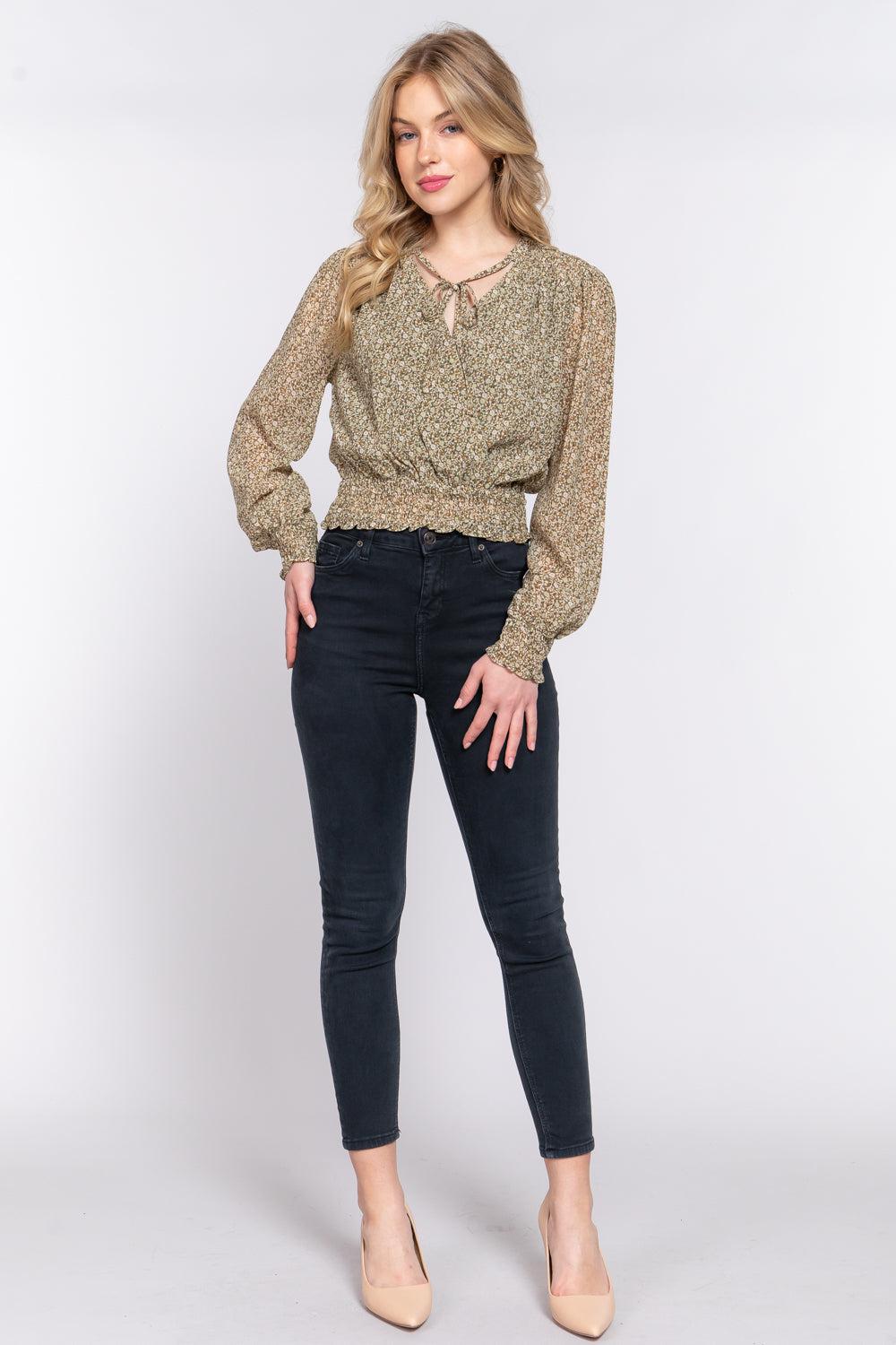 Woman wearing olive floral smocked chiffon blouse with long sleeves and surplice tie detail, paired with black jeans and nude heels.