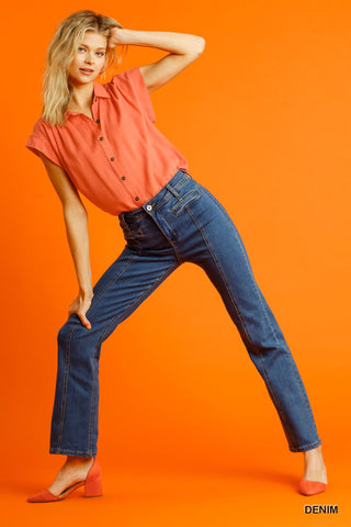 Woman wearing straight cut denim jeans with pockets and a coral blouse, posing against an orange background.