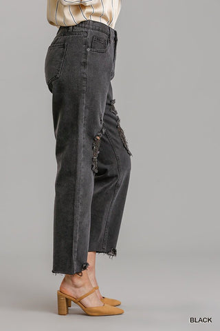 Black straight cut distressed denim jeans with raw hem featuring 5 pockets, shown from the side worn by a model in heels.