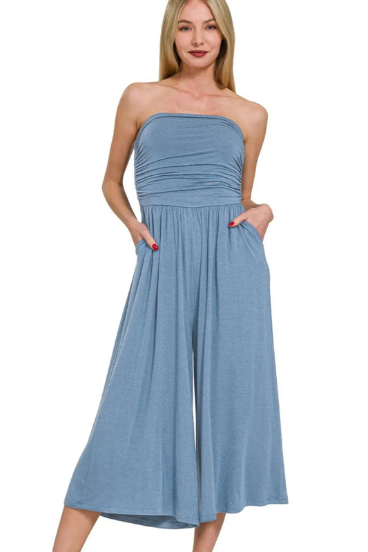 Woman in blue strapless midi dress with ruched bodice and side pockets, standing against white background with hands in pockets.