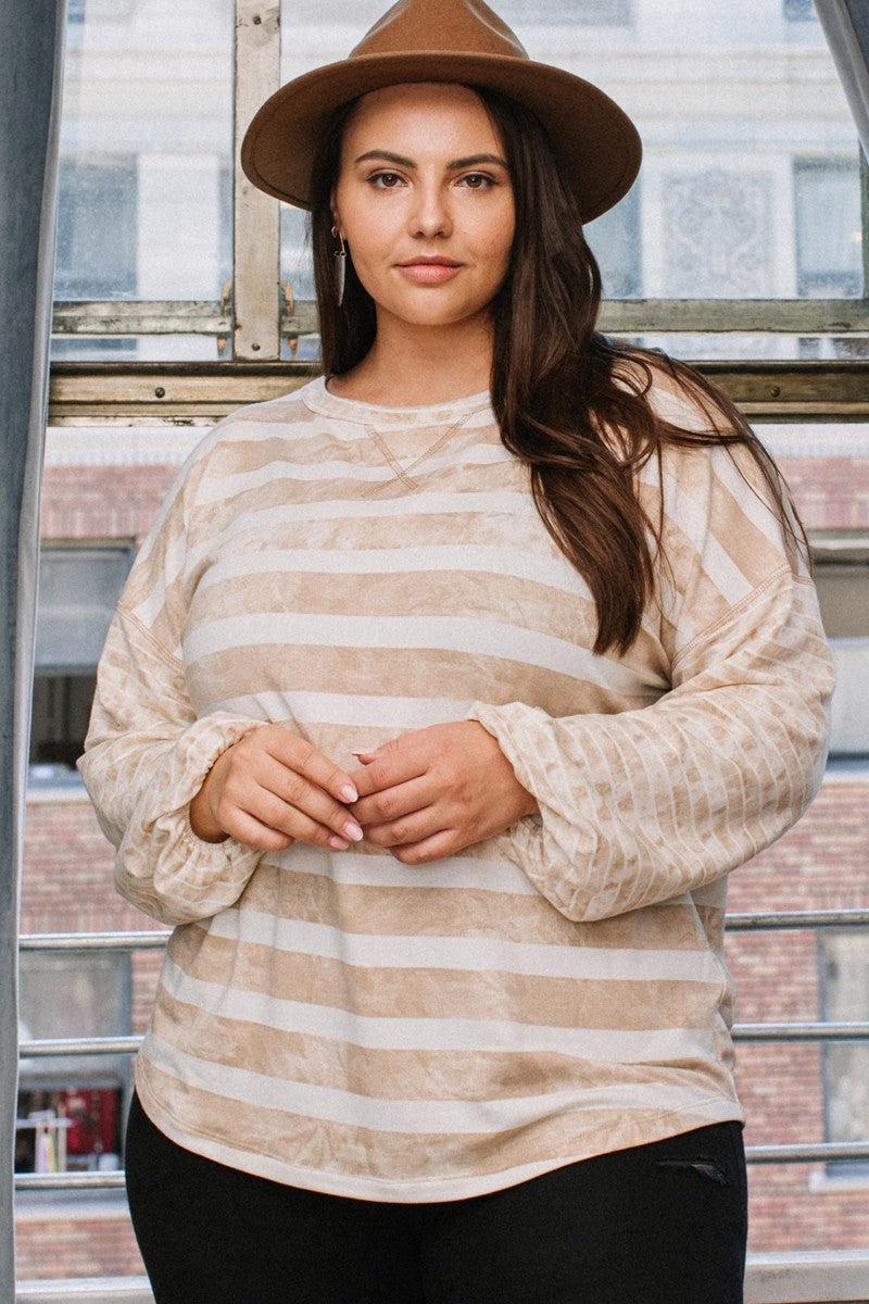 Plus-size model wearing a taupe stripe printed French terry top with a round neckline and contrast long sleeves, styled with a brown hat.