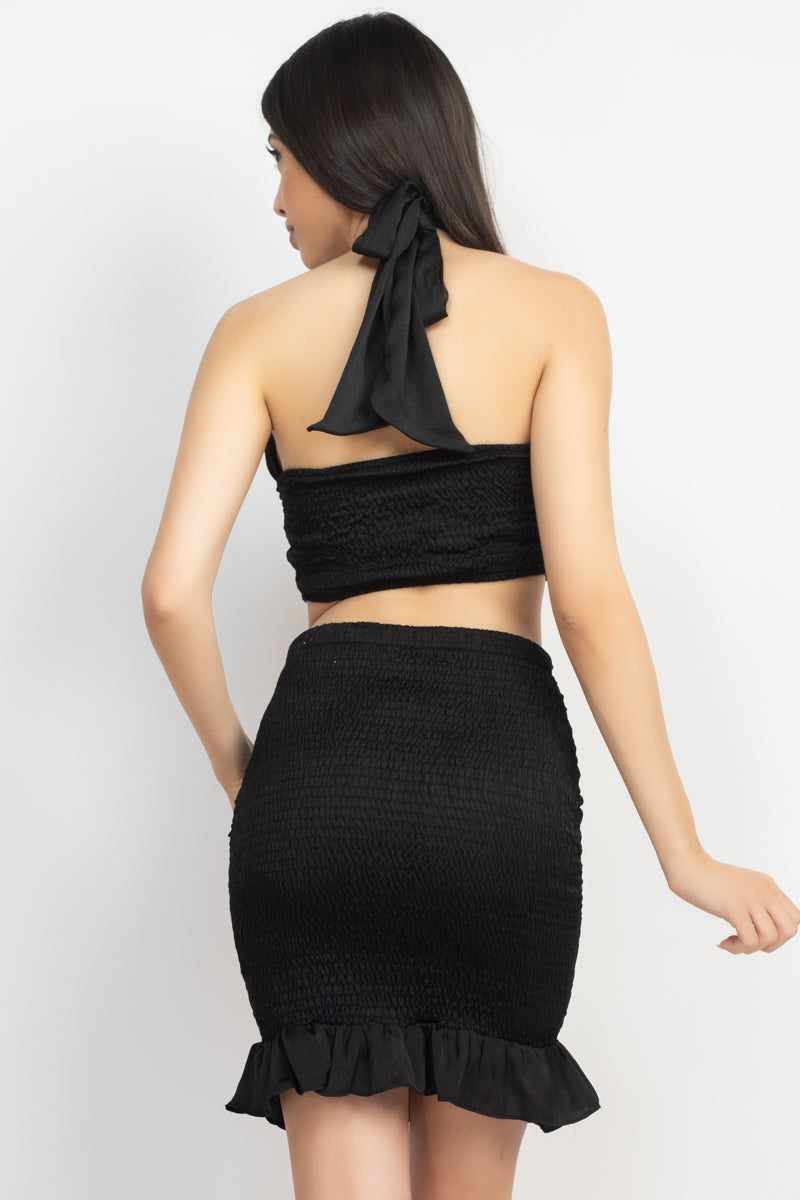 Black halter neck crop top and skirt set with shirred and smocking details, featuring a ruffled hem and open back design.