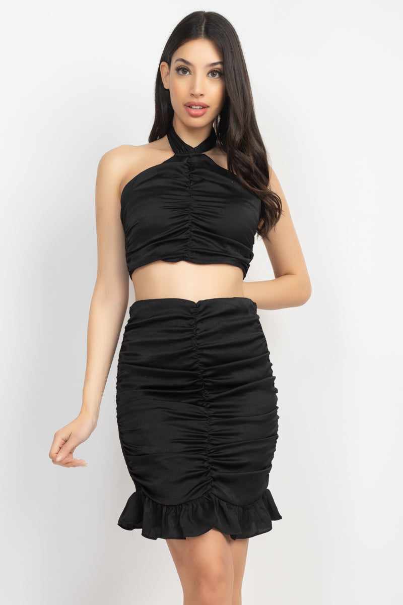 Black halter neck crop top and mini skirt set with shirred and smocking design, featuring a ruffled hem and open back. 100% polyester.