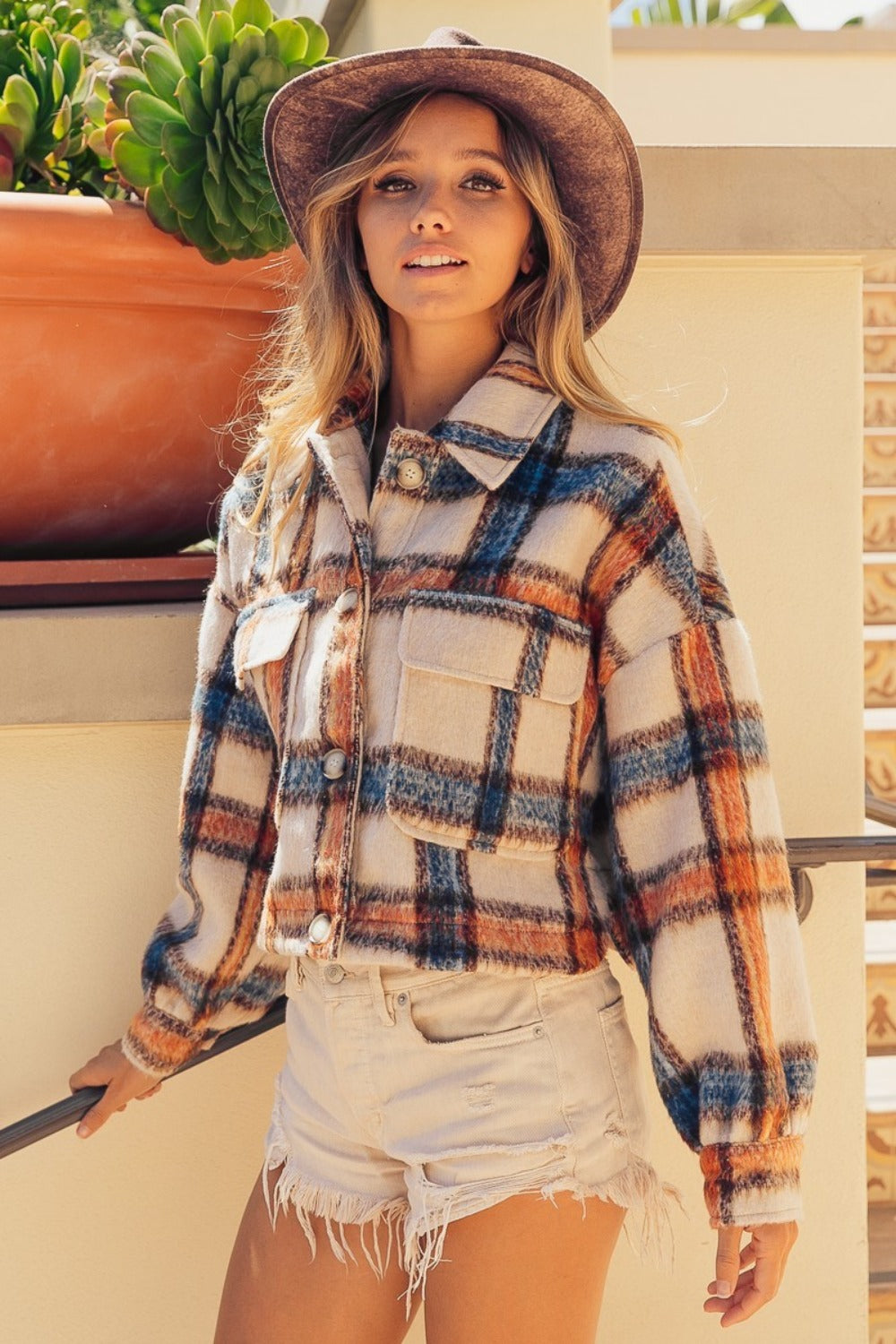 Woman in ivory multi-color brushed plaid crop jacket with pockets, wearing a hat and shorts, standing by a plant.