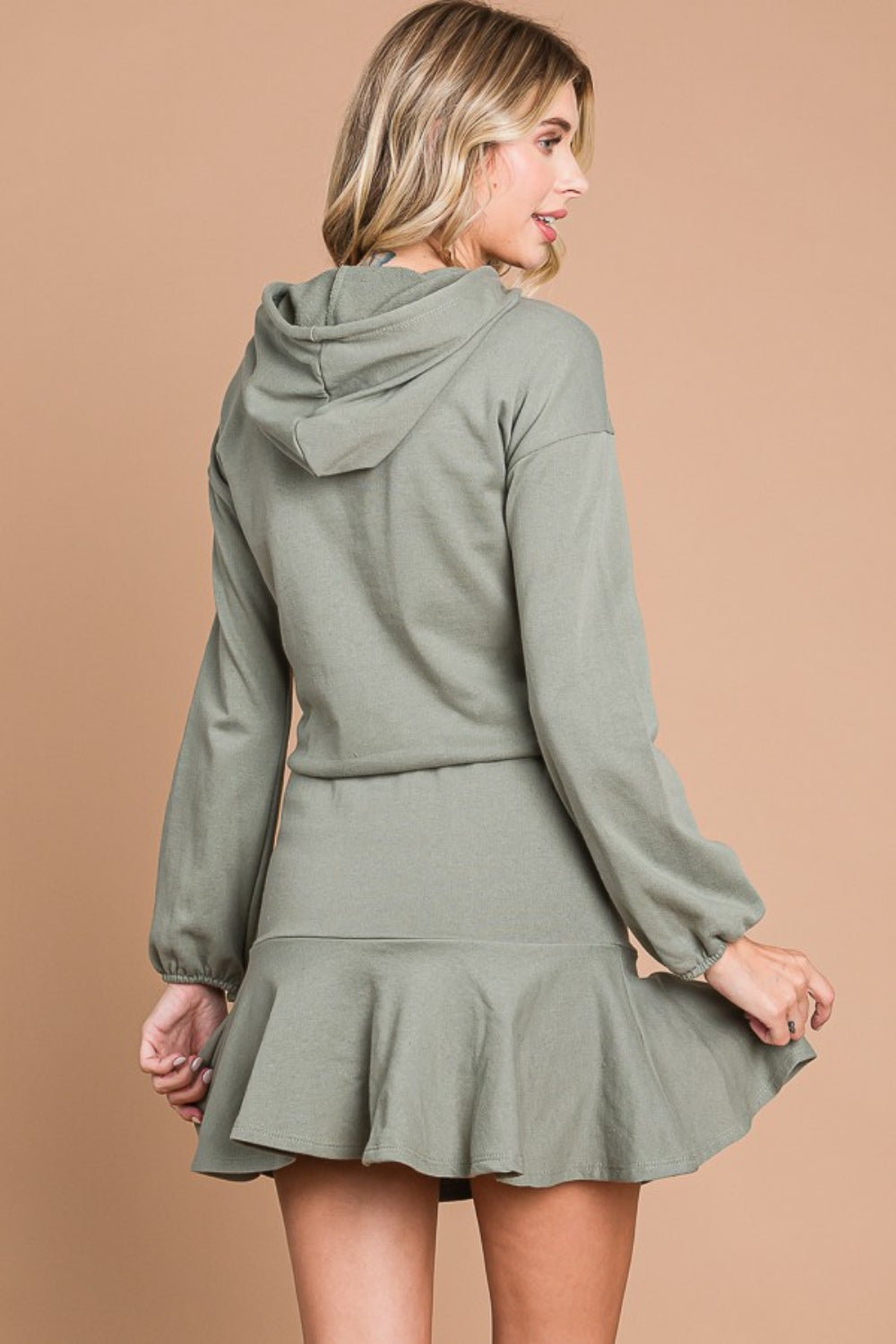 Woman wearing olive drawstring dropped shoulder hooded mini dress with ruffled hem and pockets, made of cotton-polyester blend.