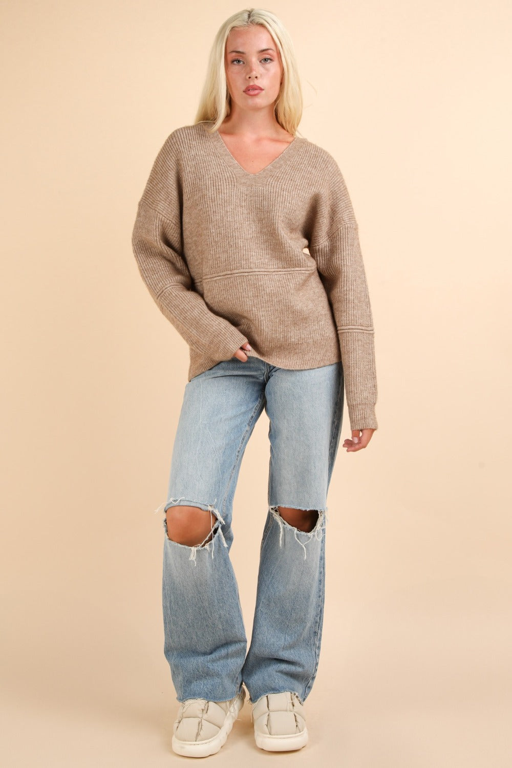 Woman wearing a beige V-neck sweater with drop shoulders and blue ripped jeans, showcasing a casual stylish outfit.