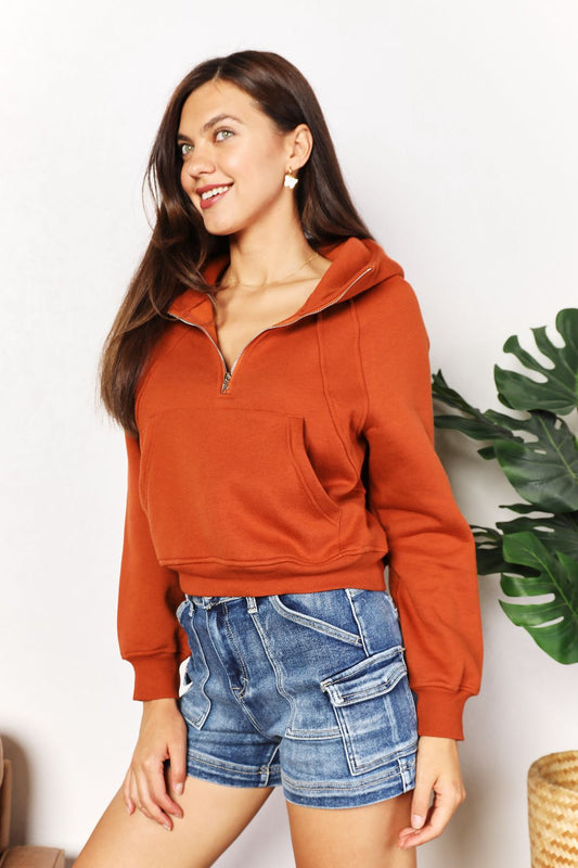 Woman wearing orange half-zip long sleeve hoodie with collared neck and cropped length, paired with denim shorts, chic style.