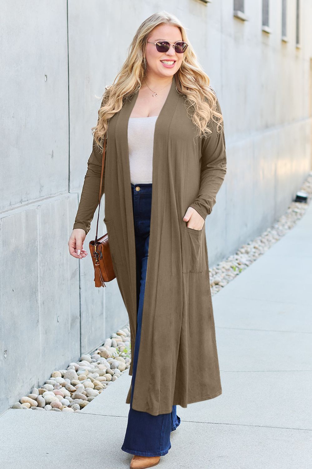 Woman wearing open front long sleeve cover up with pockets, paired with jeans and sunglasses, on a city street.