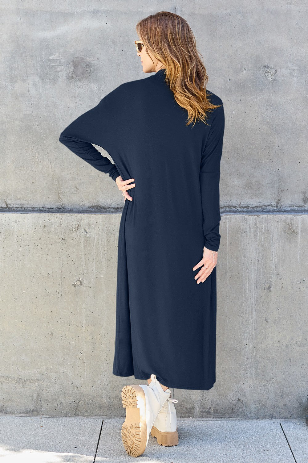 Woman wearing open front long sleeve cover up with pockets, slightly stretchy fabric, navy color, back view against concrete wall.