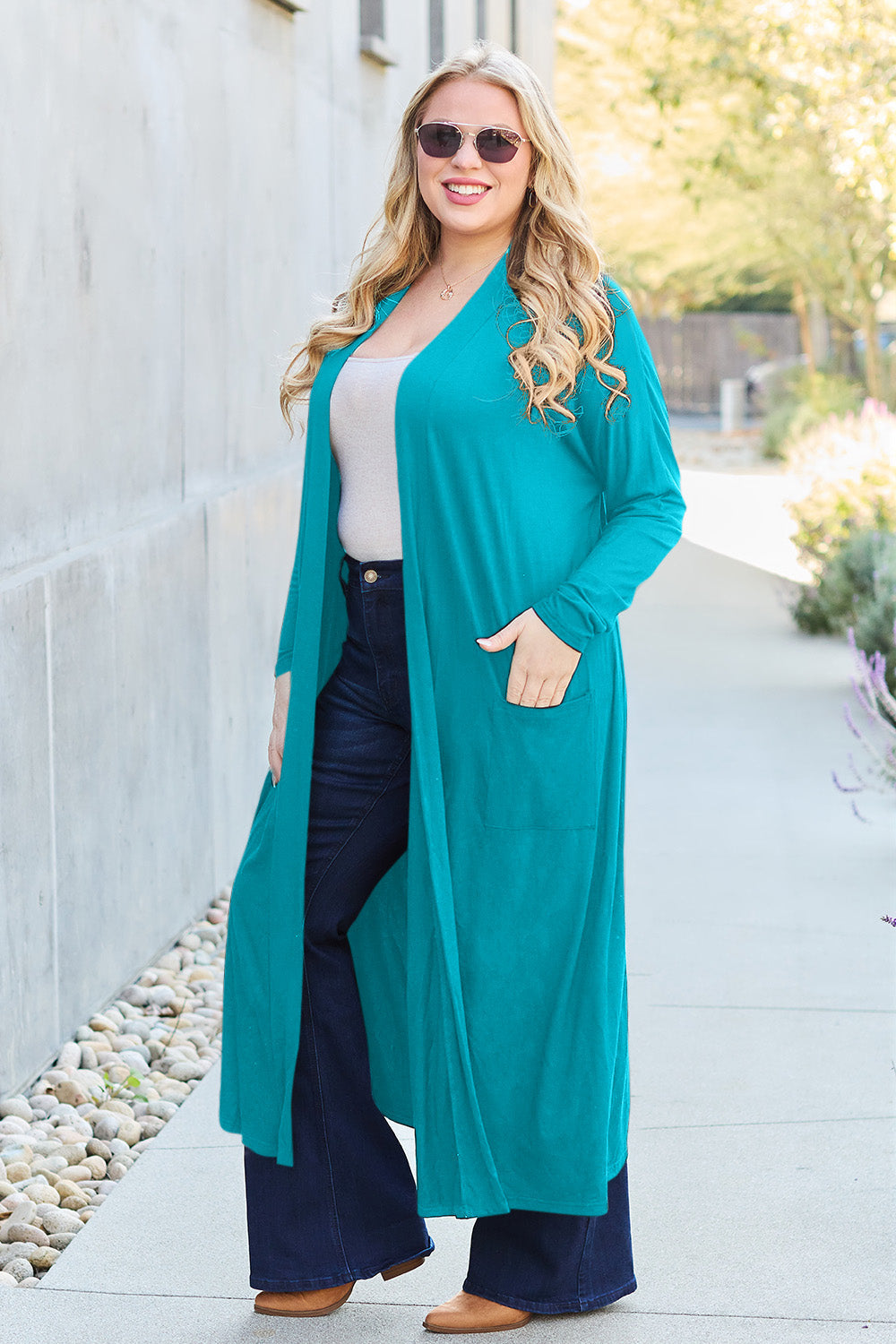 Woman wearing teal open front long sleeve cover up with pockets, paired with jeans and sunglasses, walking outdoors.