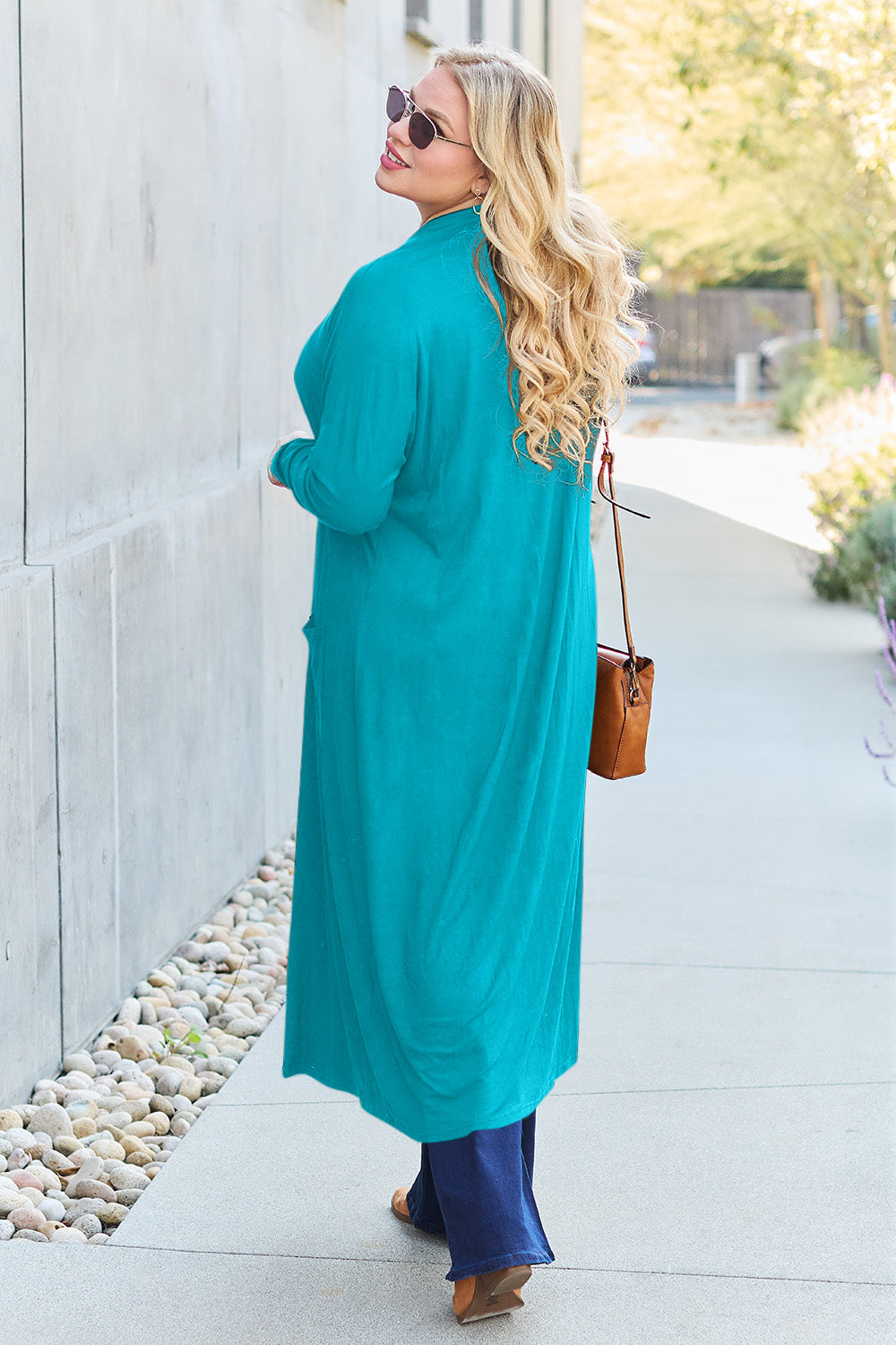 Woman wearing a turquoise open front long sleeve cover up with pockets, paired with sunglasses and carrying a brown handbag.