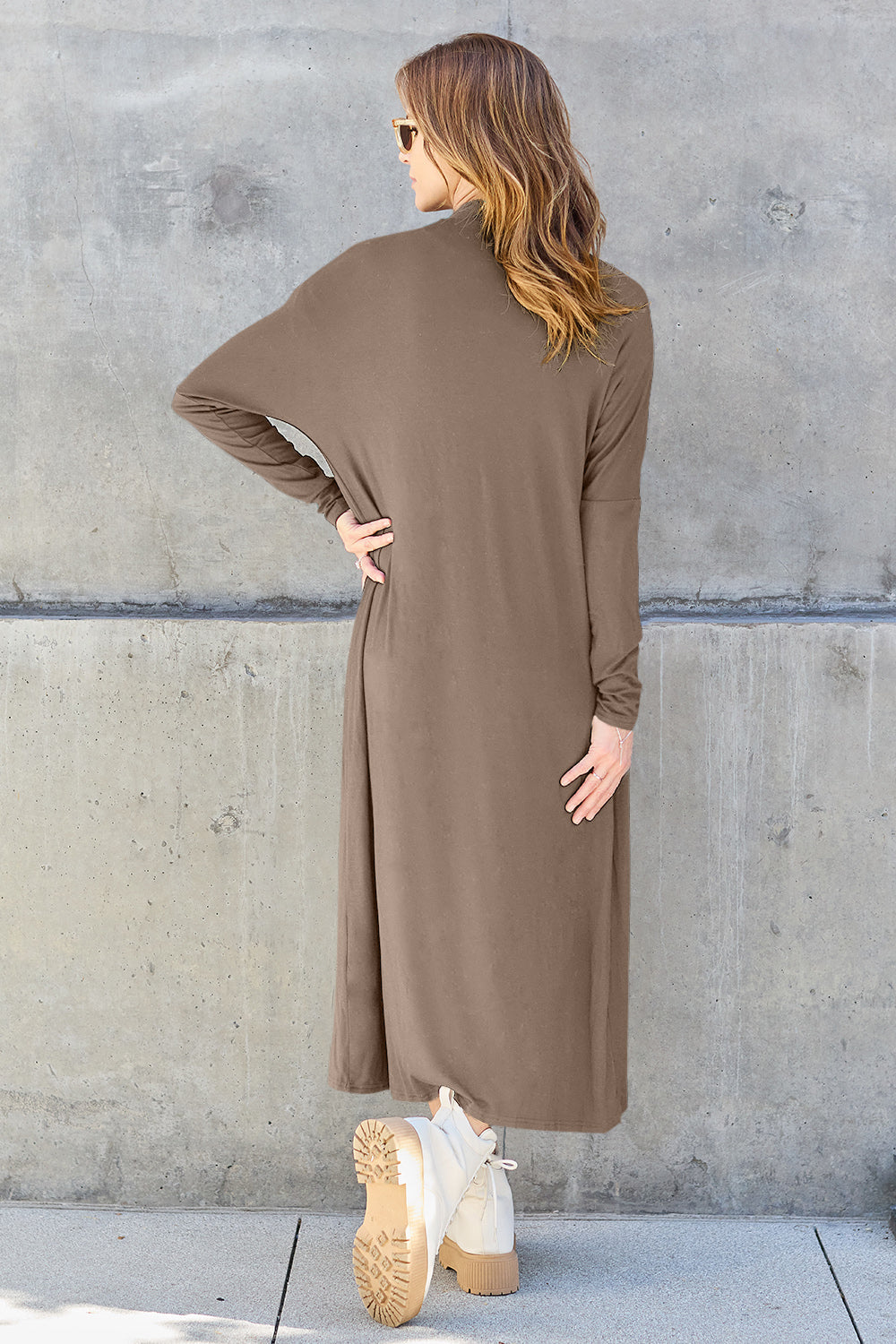 Woman wearing open front long sleeve cover up with pockets, showing back view against concrete wall.