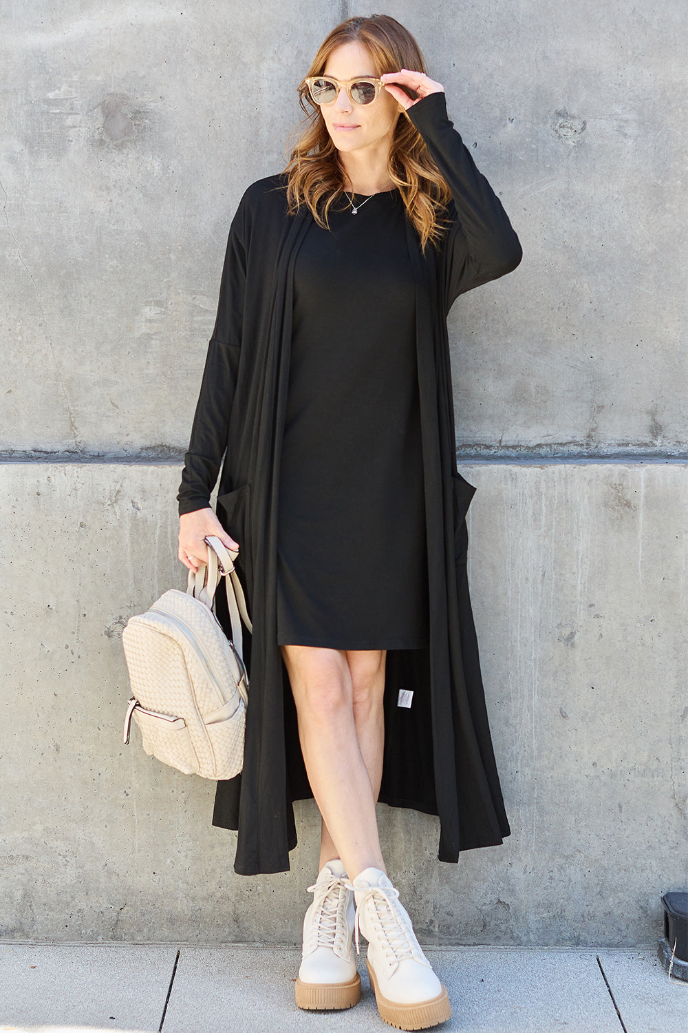 Woman wearing a black open front long sleeve cover up with pockets, paired with a black dress, white boots, and carrying a beige backpack.