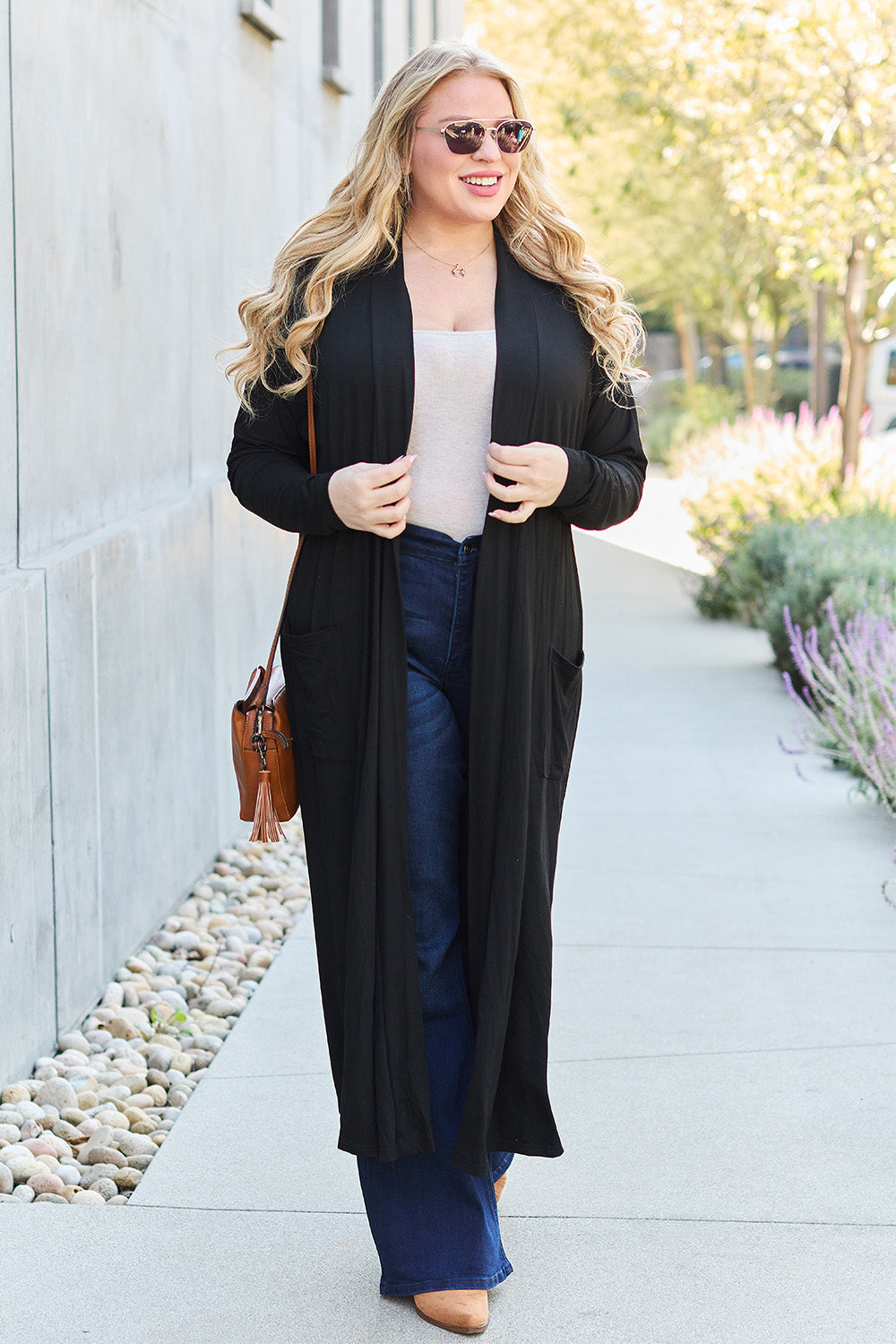 Woman wearing an open front long sleeve cover-up with pockets, paired with jeans and sunglasses, walking on a sunny day.