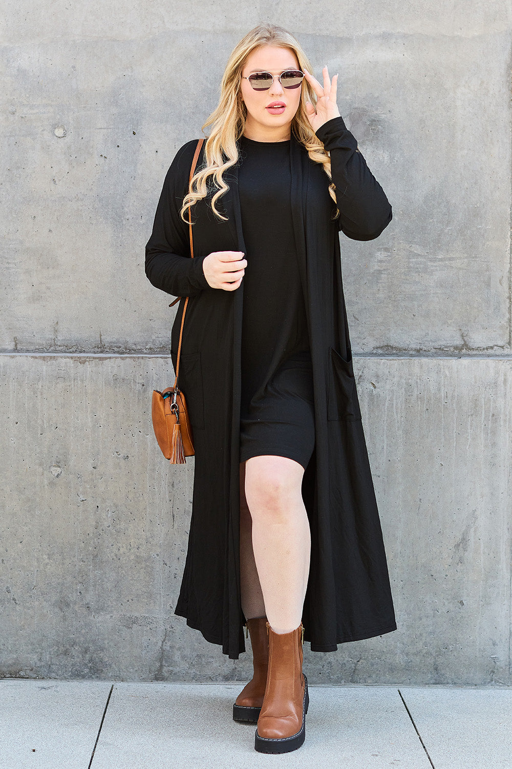 Woman wearing black open front long sleeve cover up with pockets, paired with brown boots and sunglasses, posing outdoors.