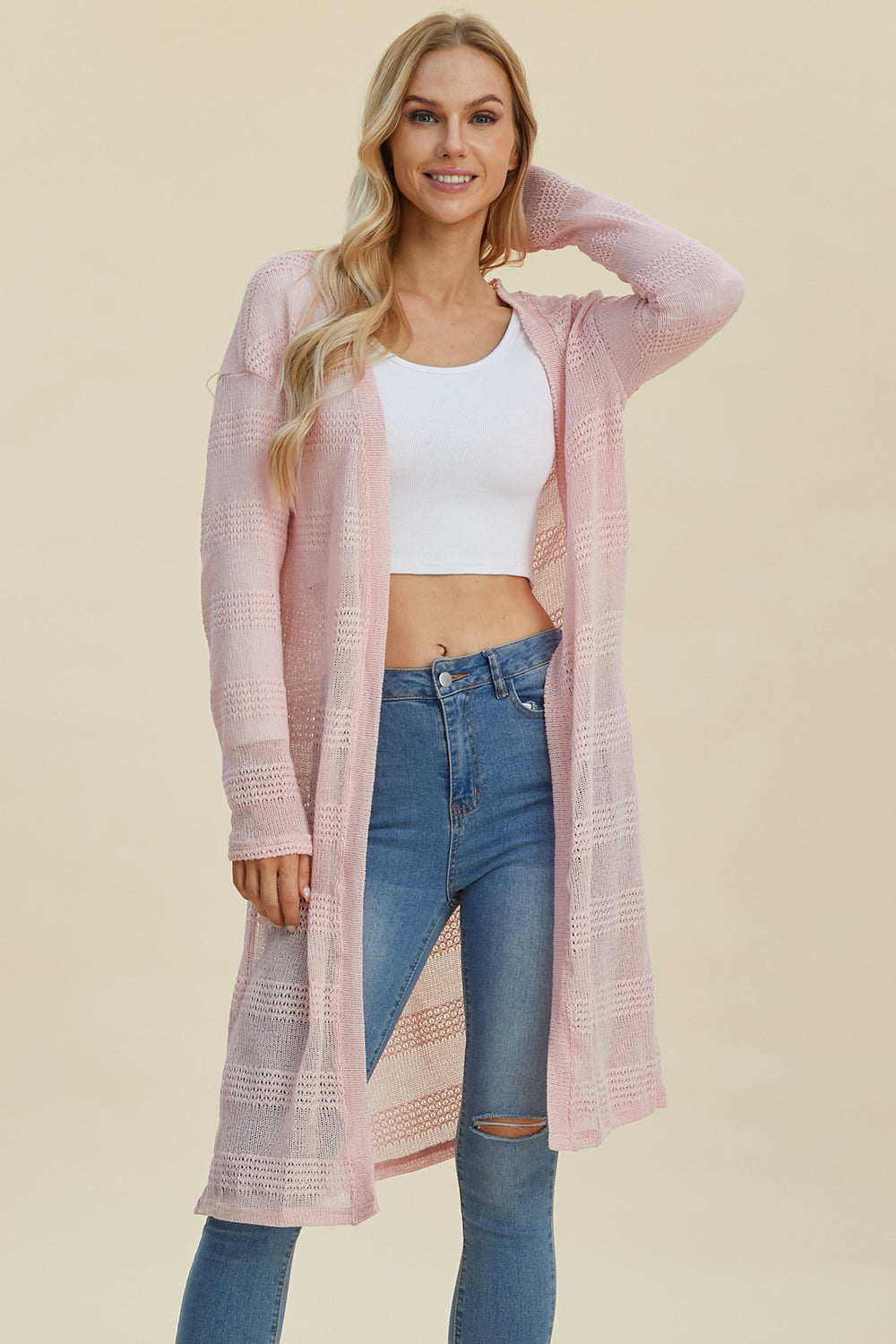 Woman wearing open front longline cardigan in pink with openwork design over white top and jeans.