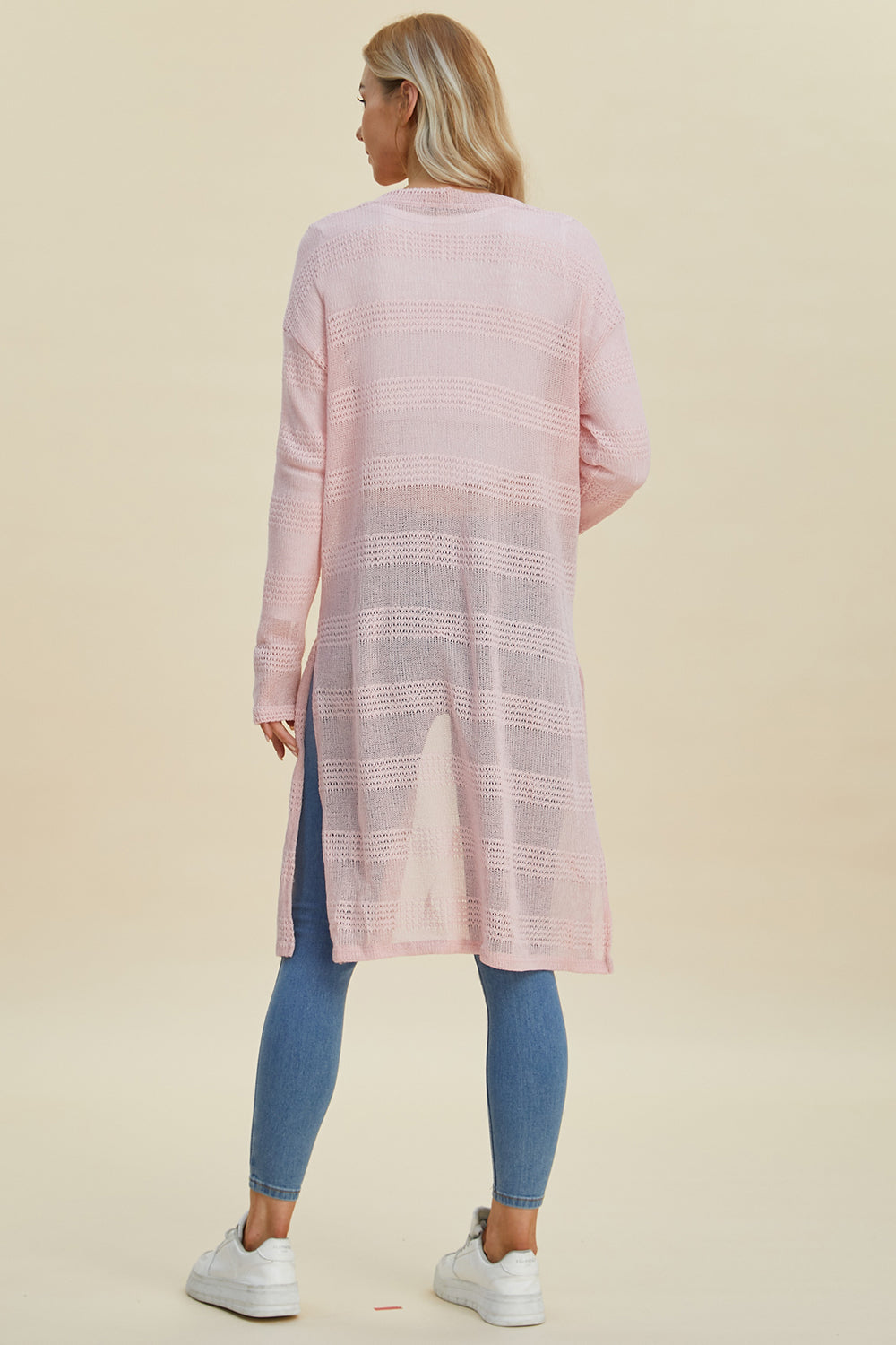 Woman wearing pink open front longline cardigan with openwork design, paired with blue jeans and white sneakers, viewed from the back.