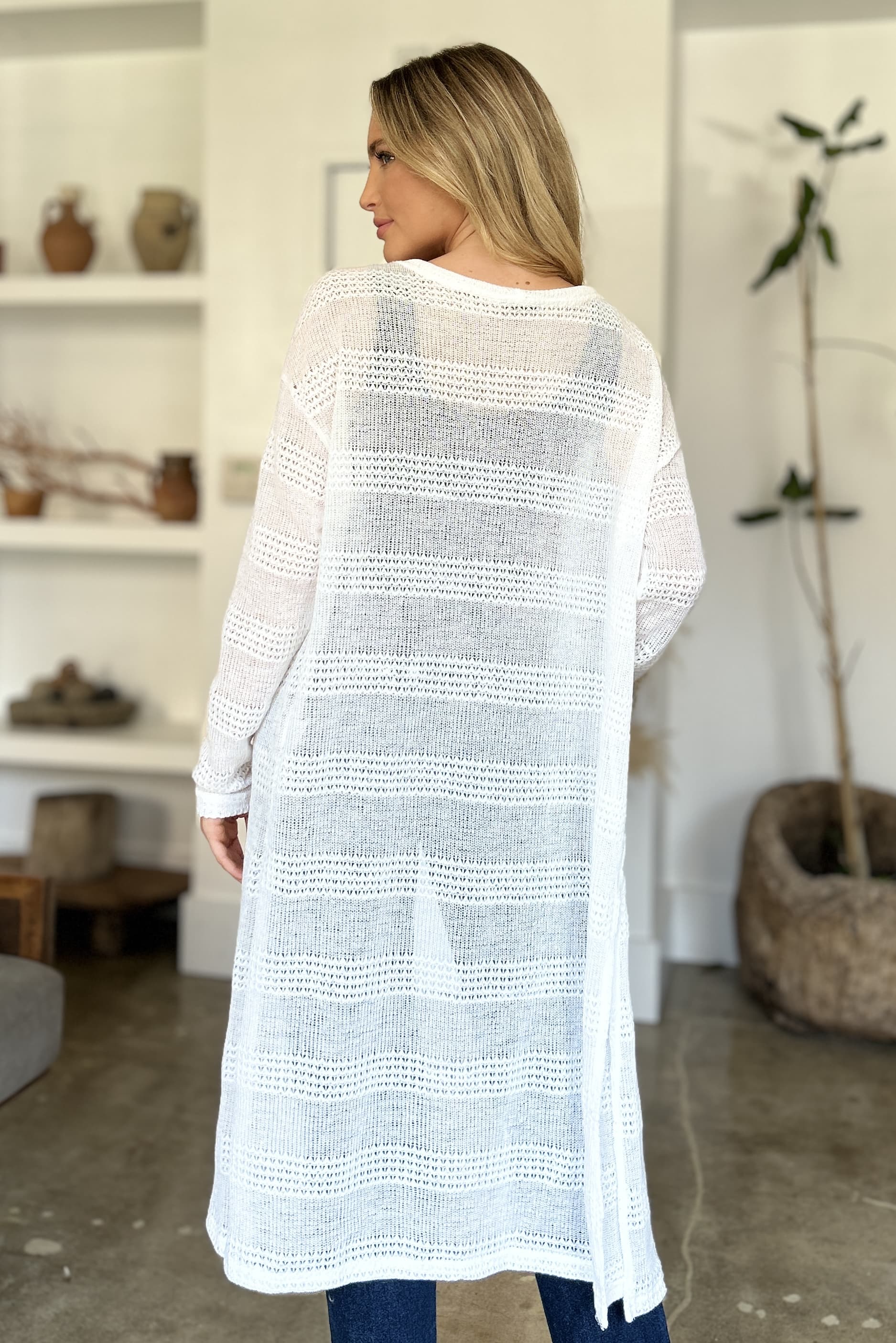 Woman wearing open front longline cardigan with openwork design, showcasing moderate stretch and polyester material in a stylish room.