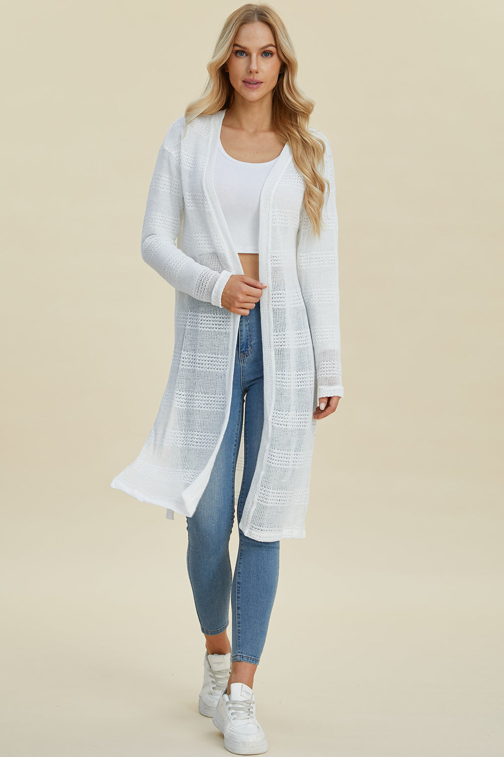 Woman wearing open front longline cardigan with openwork design, moderate stretch, made of 100% polyester, styled with jeans and sneakers.