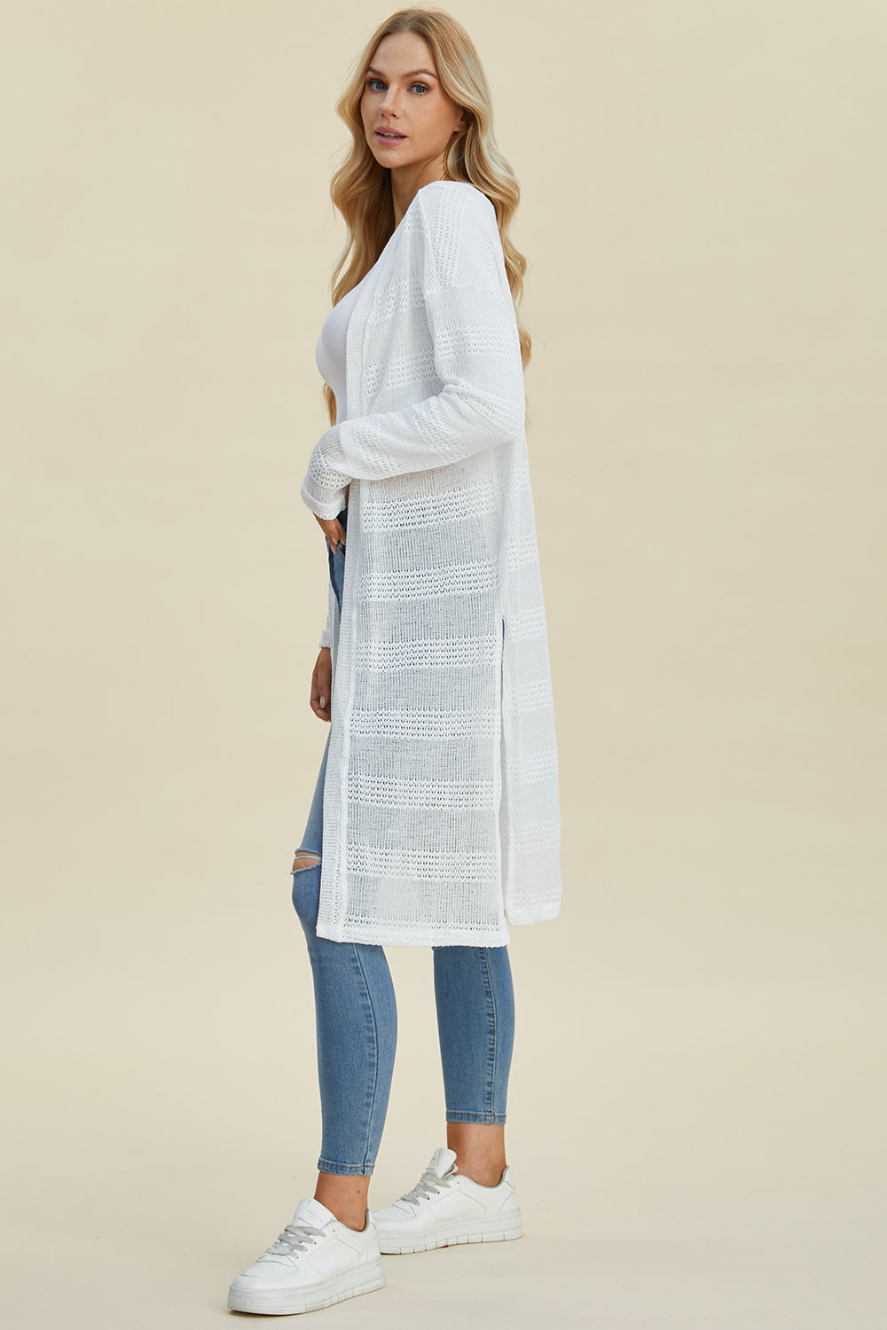 Woman wearing a white open front longline cardigan with openwork design and moderate stretch, paired with jeans and sneakers