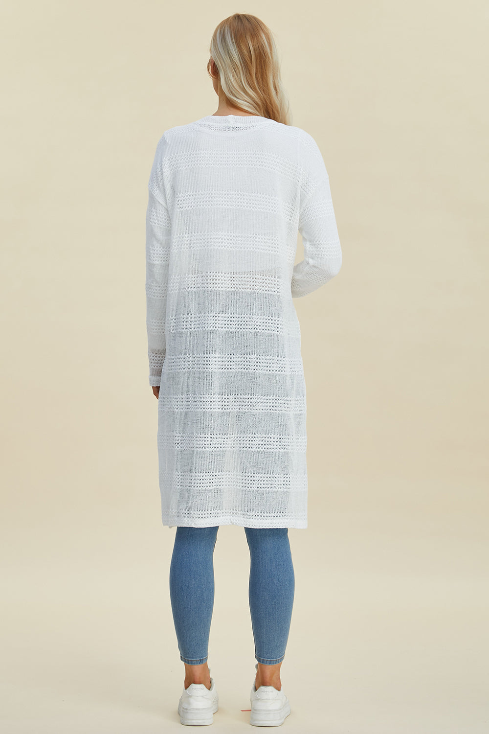 Woman wearing white openwork longline cardigan with moderate stretch and full size, paired with blue jeans and white sneakers, back view.