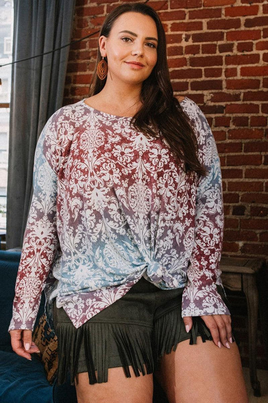 Woman wearing a printed long-sleeve top with knot detail and fringe shorts, standing in a stylish room with brick wall background.