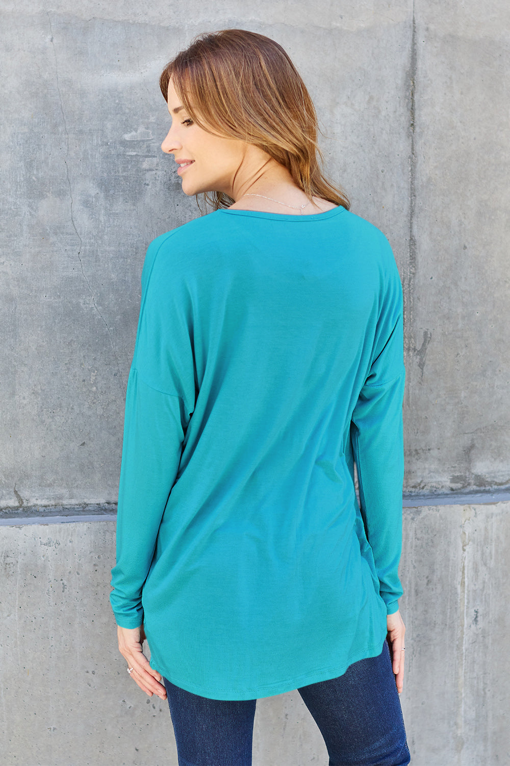 Woman wearing teal round neck dropped shoulder t-shirt, back view in front of a concrete wall.