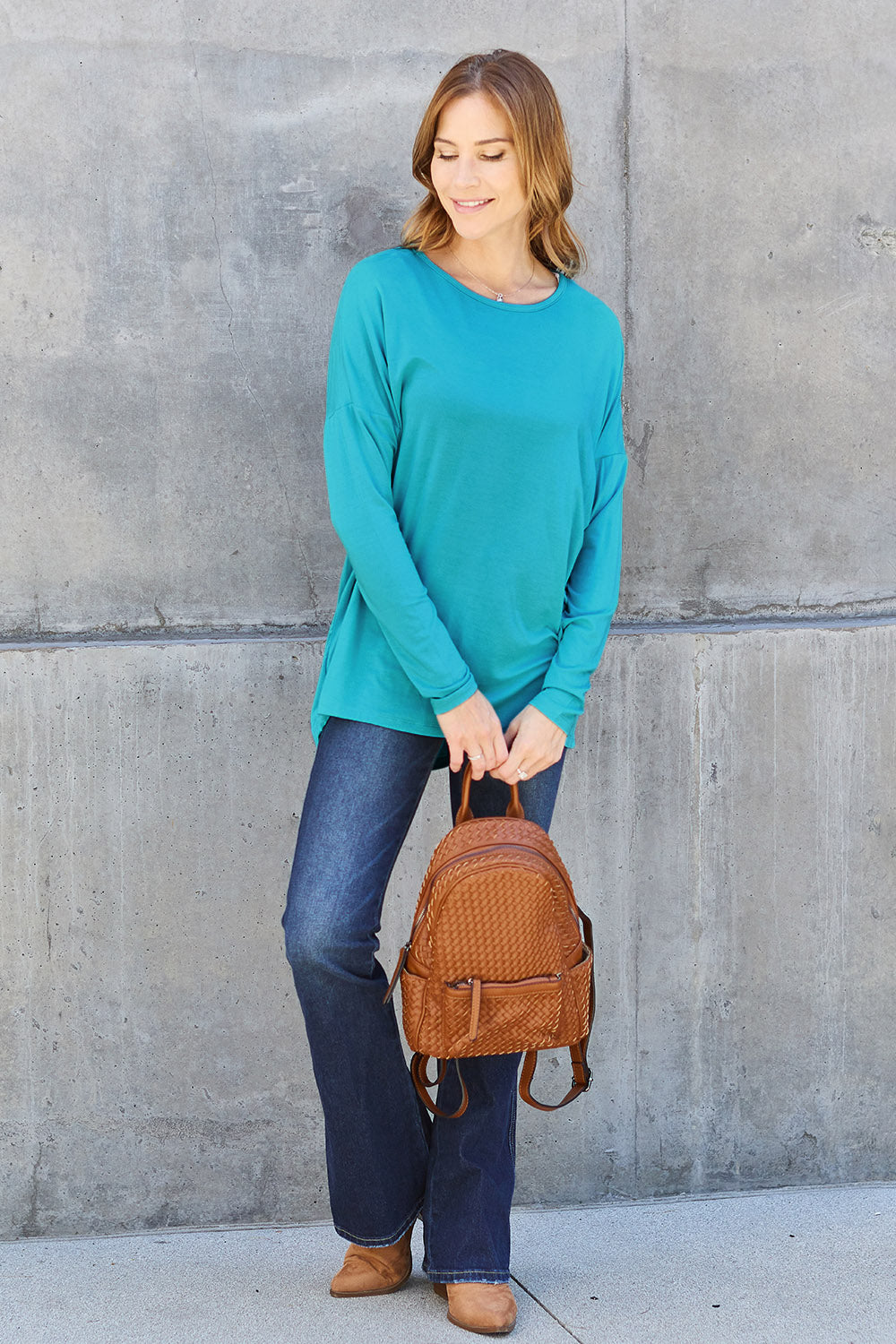 Woman wearing a round neck dropped shoulder t-shirt in teal, paired with jeans and holding a brown backpack.