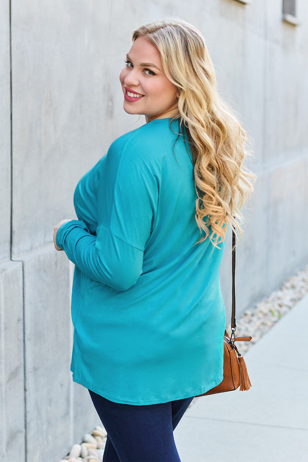 Woman wearing a turquoise round neck dropped shoulder T-shirt with long sleeves, paired with dark jeans and a crossbody bag outdoors.