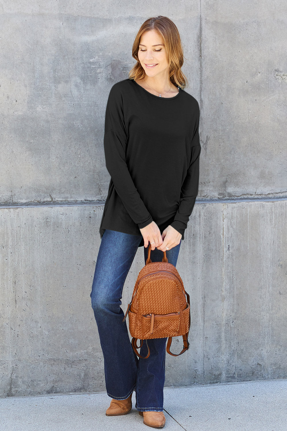 Woman in black round neck dropped shoulder t-shirt with jeans and brown backpack standing against a concrete wall.