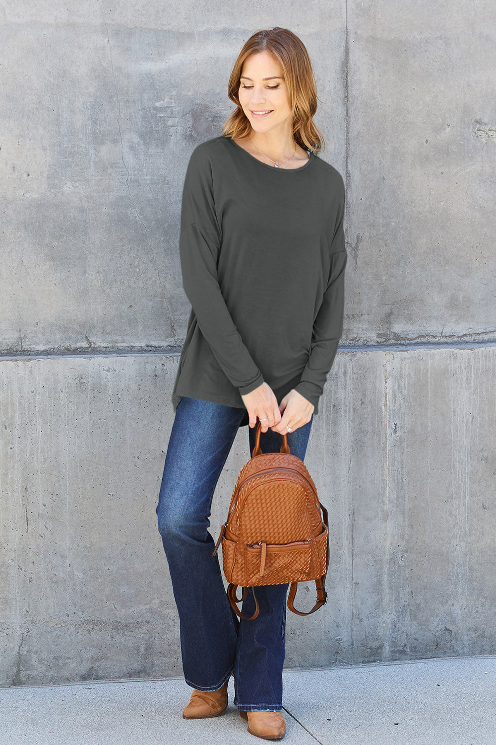 Woman wearing a round neck dropped shoulder t-shirt in dark green, paired with jeans and a brown backpack, standing against a concrete wall.