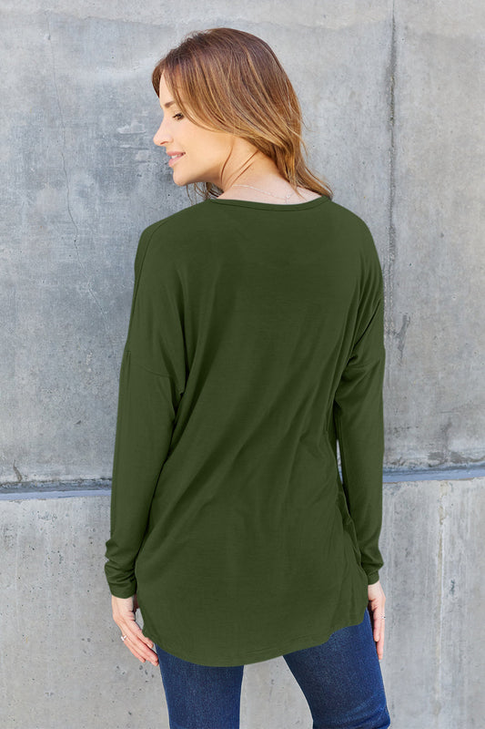 Woman wearing a green round neck dropped shoulder t-shirt with slightly stretchy material, made of rayon and spandex, standing against a concrete wall.