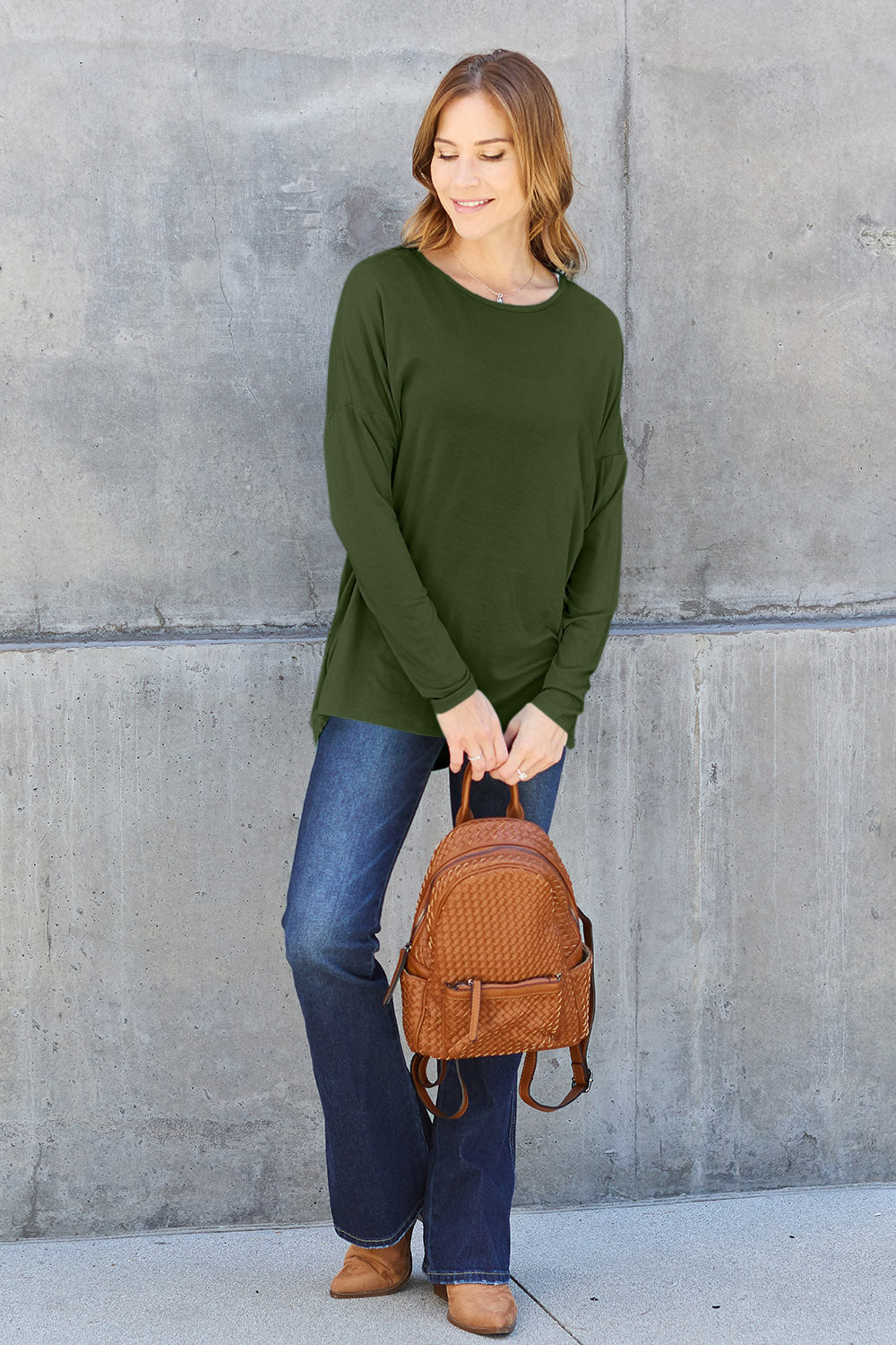 Woman wearing a green round neck dropped shoulder t-shirt with blue jeans and brown backpack against a gray wall.