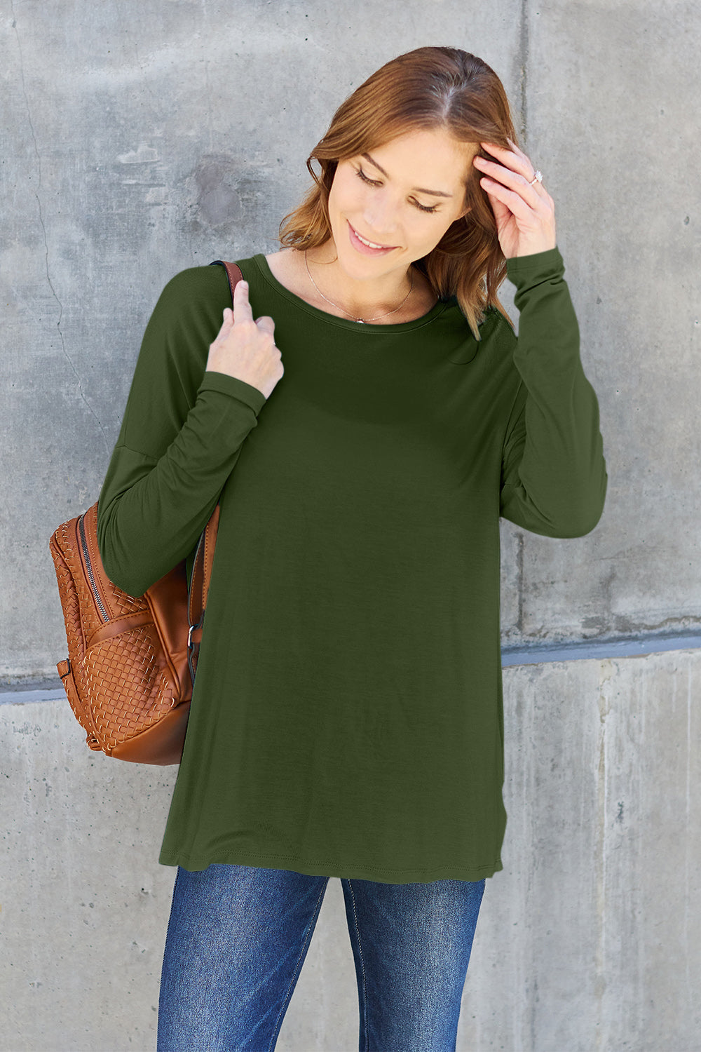 Woman wearing a green round neck dropped shoulder t-shirt paired with jeans and a brown backpack, standing against a concrete wall.