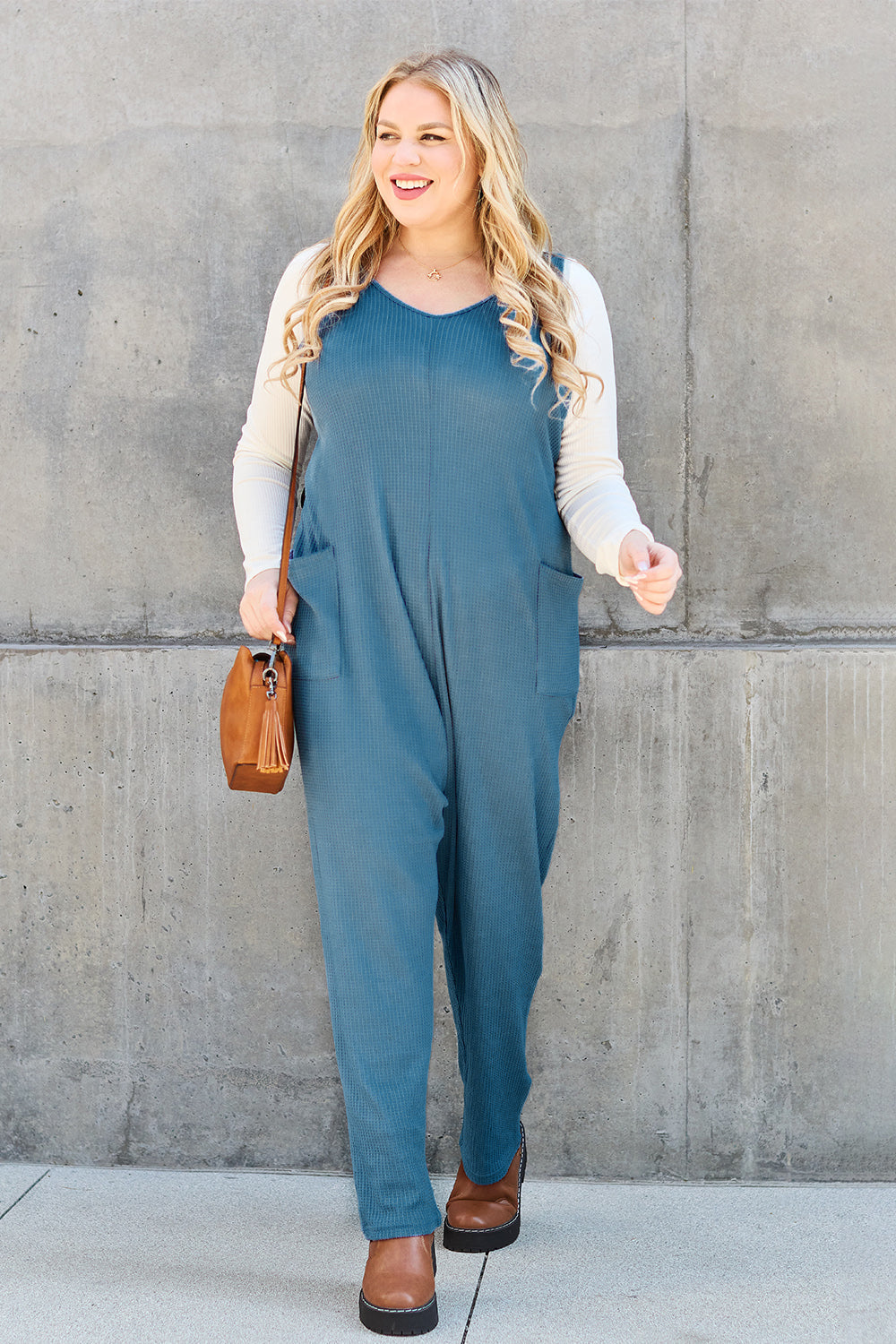 Woman wearing sleeveless straight jumpsuit with pockets, styled with a brown bag and casual shoes.