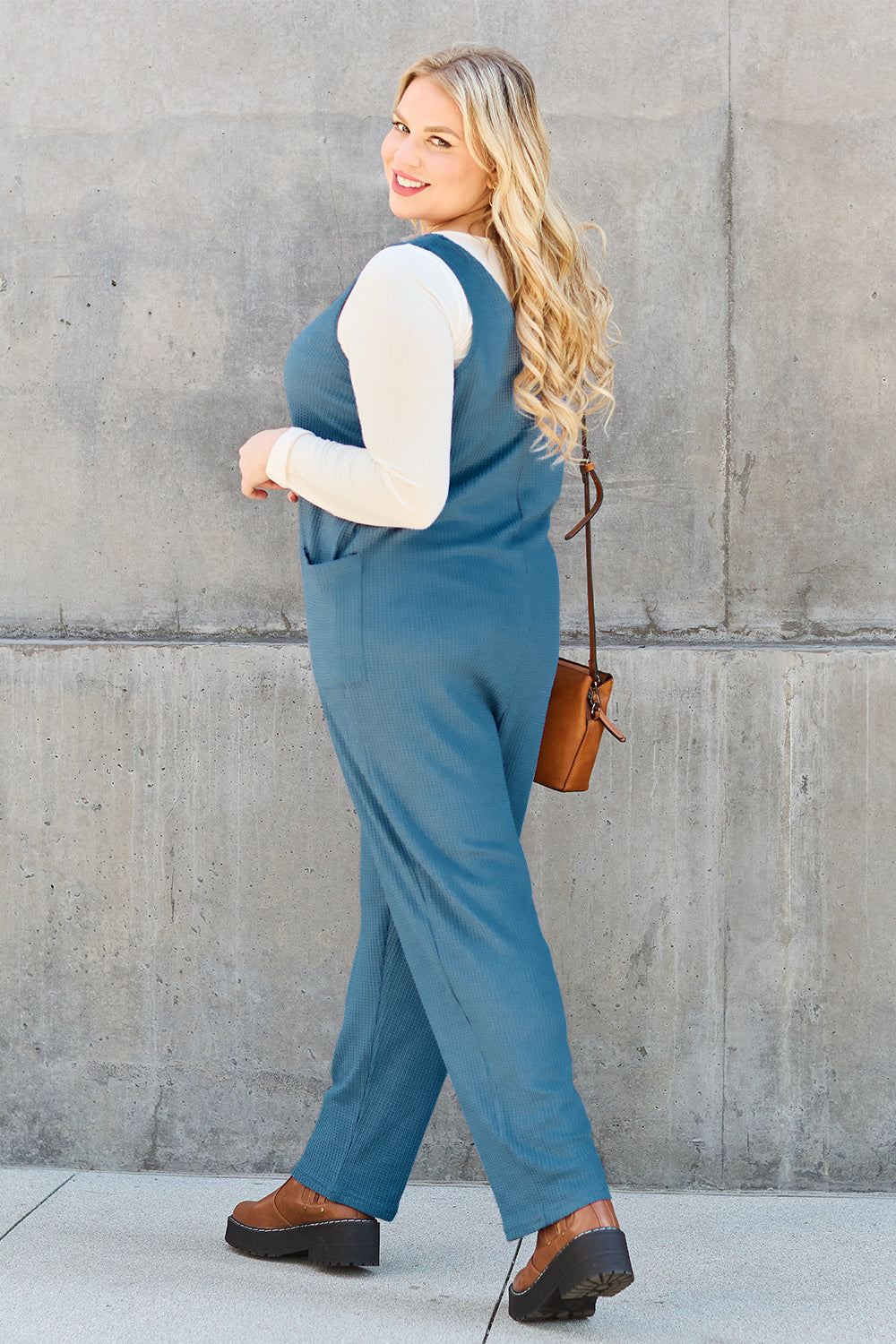 Woman wearing a sleeveless straight jumpsuit with pockets, showcasing a casual look, complemented by a small brown bag and boots.