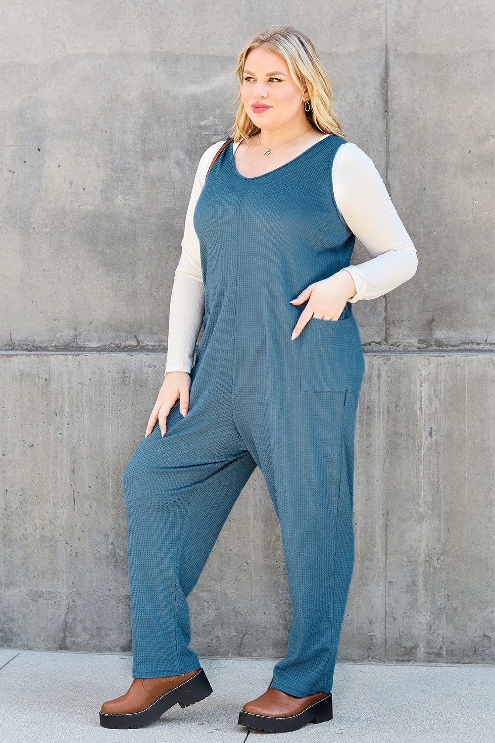 Woman wearing a sleeveless straight jumpsuit with pockets, made of 95% polyester and 5% spandex, against a concrete wall.