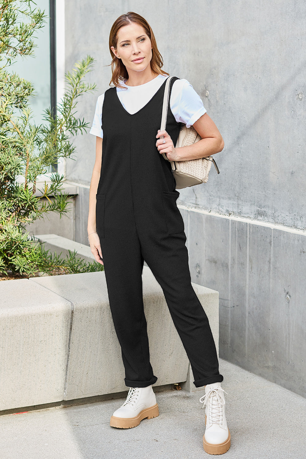 Woman wearing a sleeveless straight black jumpsuit with pockets, paired with a white shirt and boots, standing outdoors.