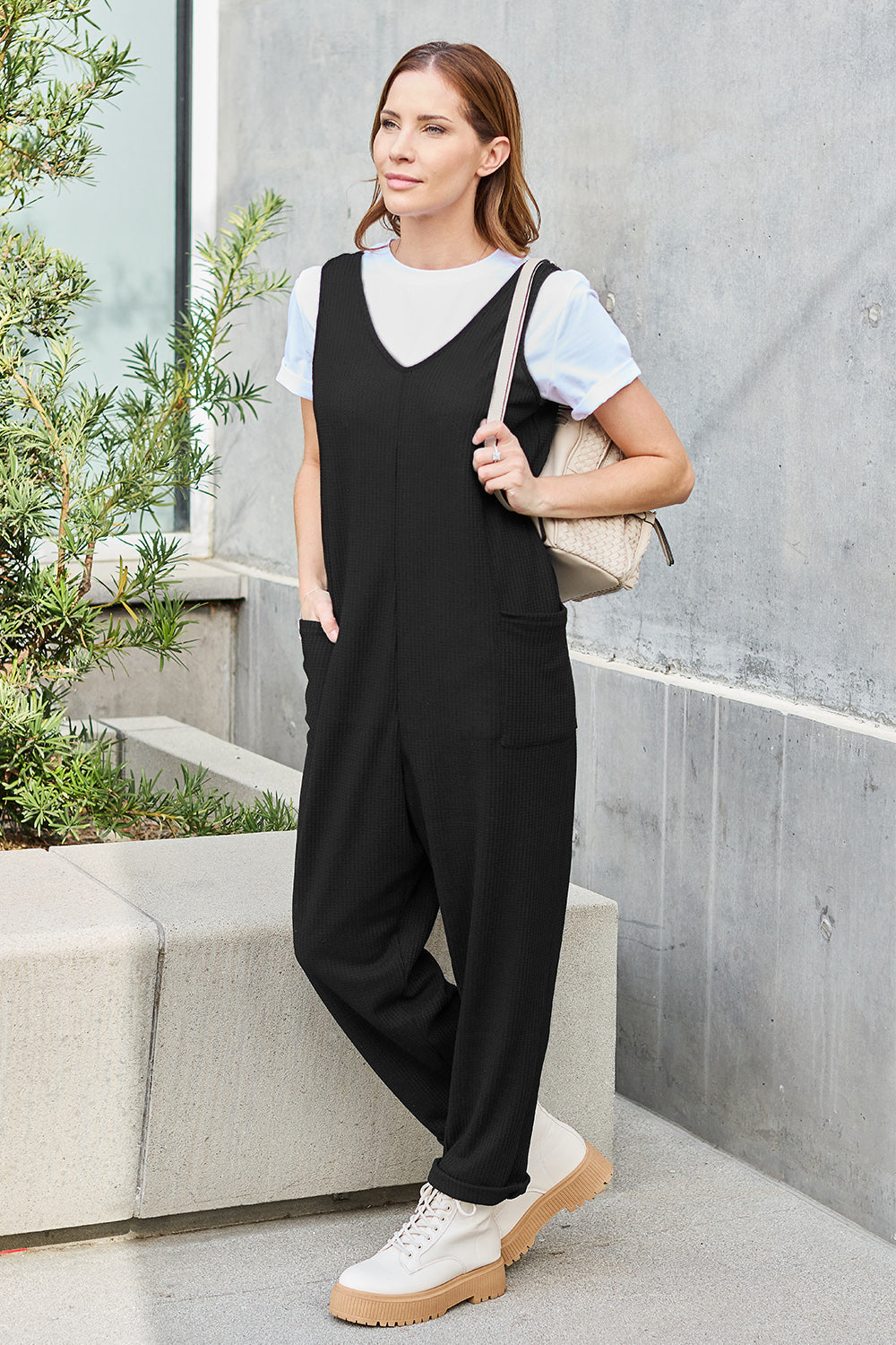 Woman wearing a sleeveless straight black jumpsuit with pockets, styled casually with a white shirt and sneakers.