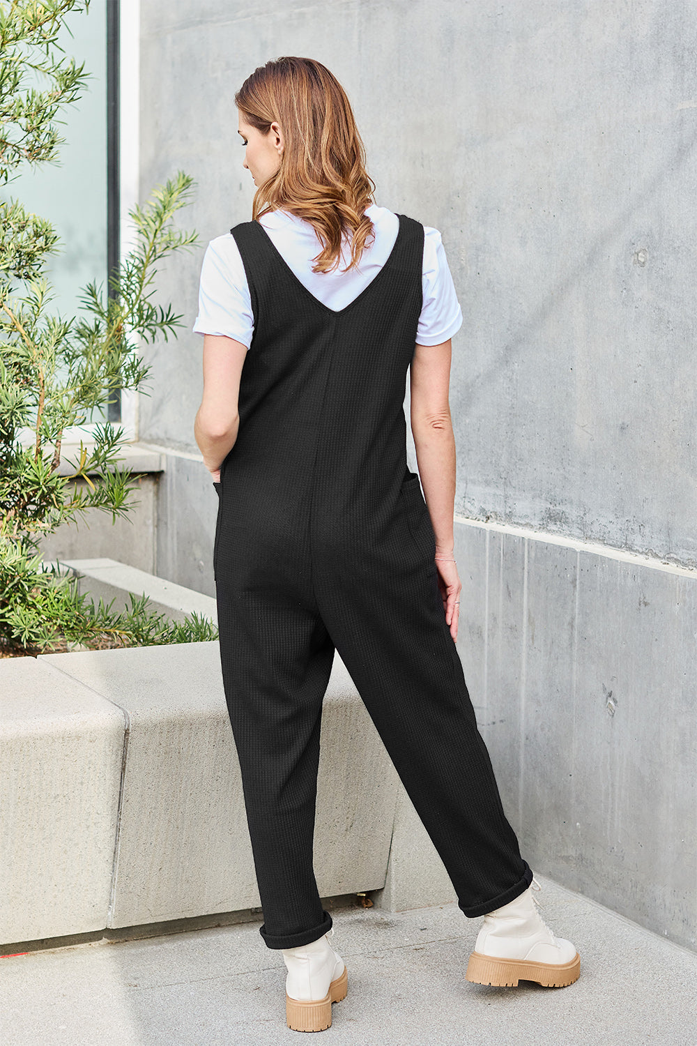 Woman wearing a black sleeveless straight jumpsuit with pocketed design, made of polyester and spandex, standing outdoors.