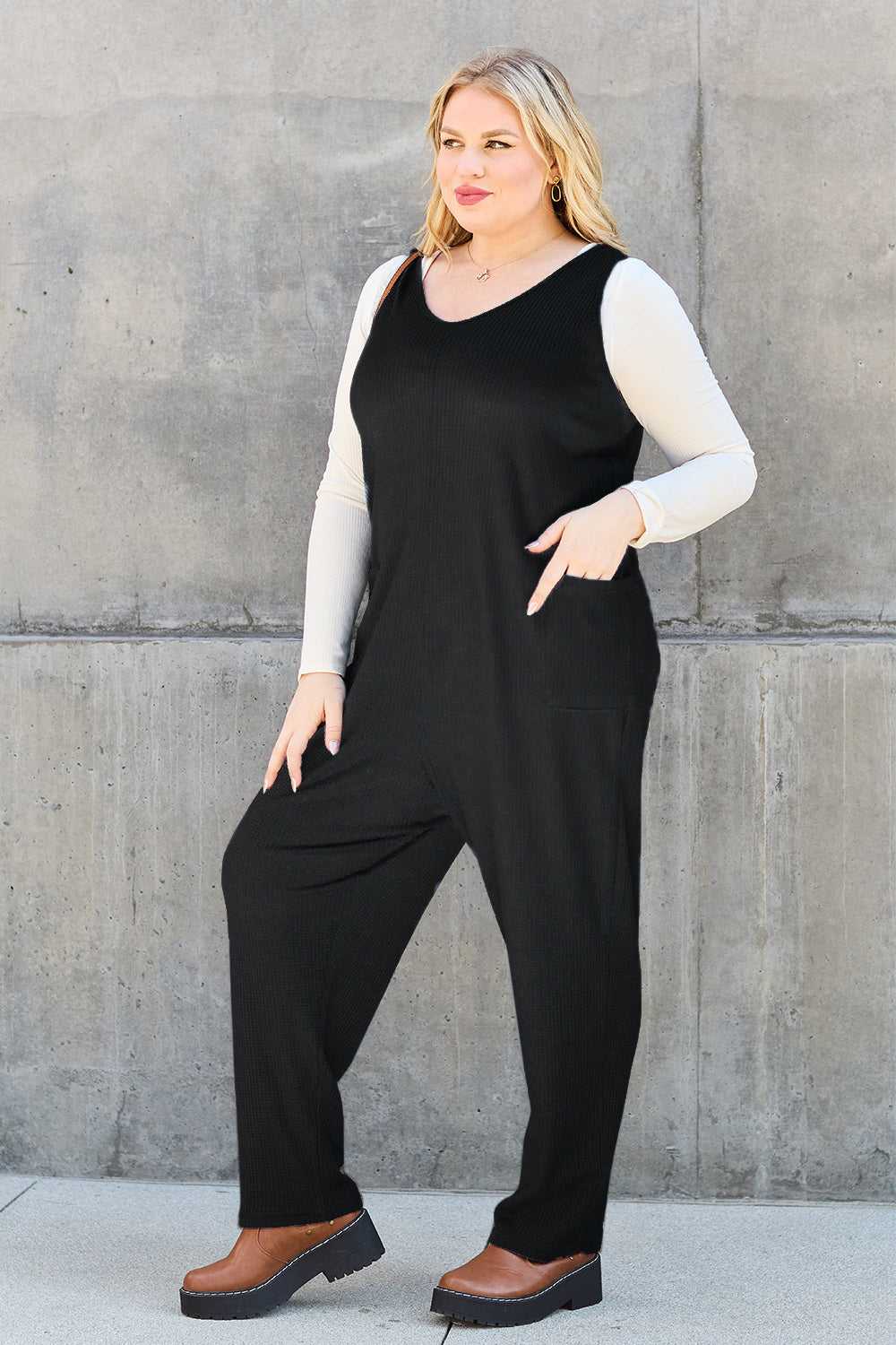 Woman wearing a sleeveless straight black jumpsuit with pockets, layered over a long-sleeve top, showcasing versatile fashion.